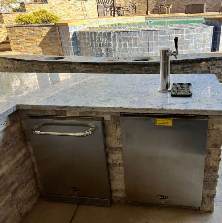 Luxury backyard kitchen and bar area featuring a built-in beer tap, dual stainless steel refrigerators, and custom stonework overlooking a residential pool.