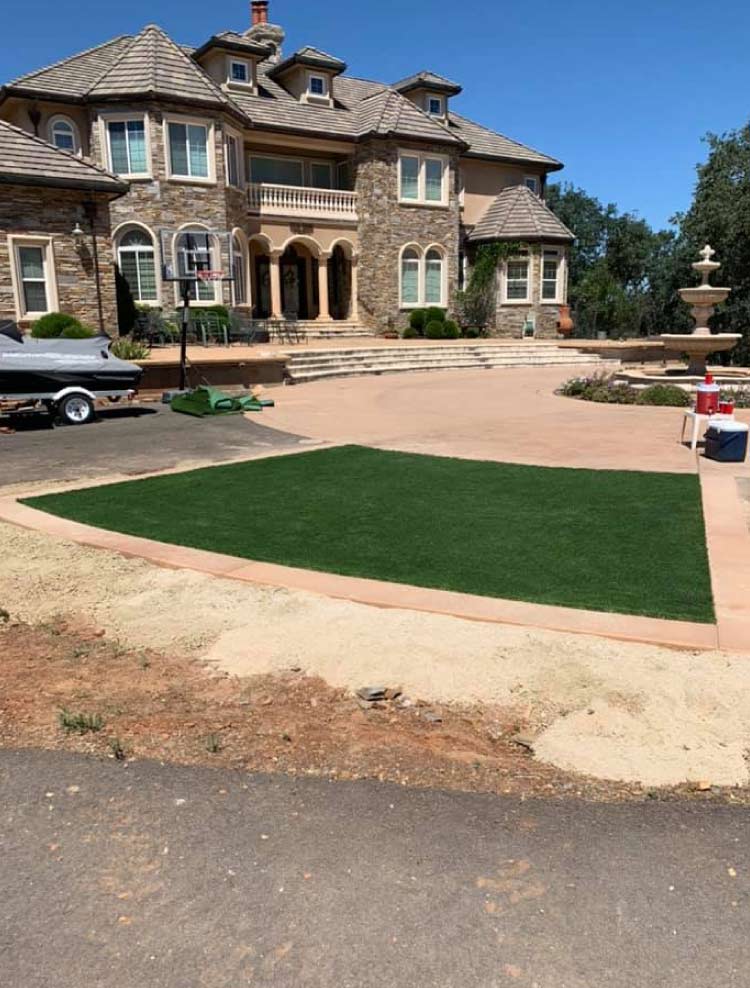 High-end front yard transformation showcasing a perfectly installed rectangular artificial turf lawn bordered by a smooth, tan concrete driveway and a large stone estate home.