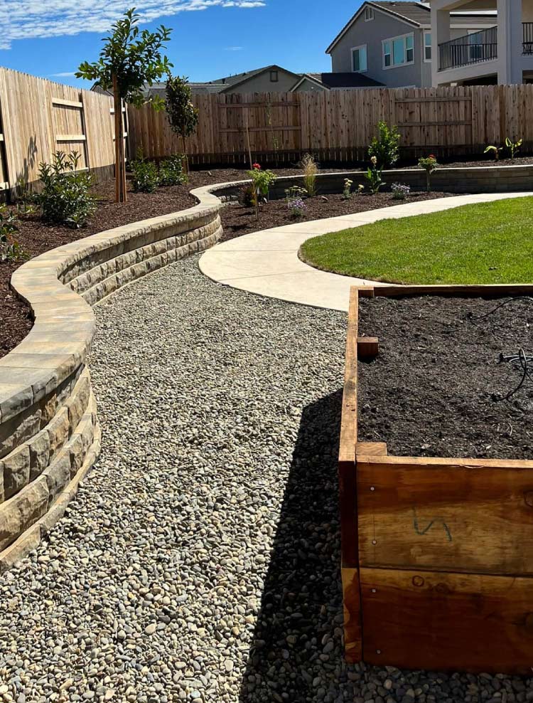 Completed backyard renovation featuring a curved multi-level stone retaining wall, a clean concrete walkway, decorative gravel, and custom wooden garden planter boxes.