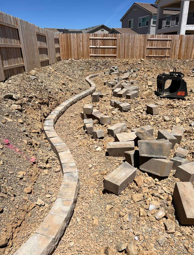 Professional backyard construction site showing the expert installation of a curved stone retaining wall foundation on a sloped residential lot.