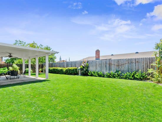 A vibrant, sun-drenched backyard featuring a perfectly manicured green lawn, a white custom patio covers with a ceiling fan, and lush privacy hedges along a wooden fence.