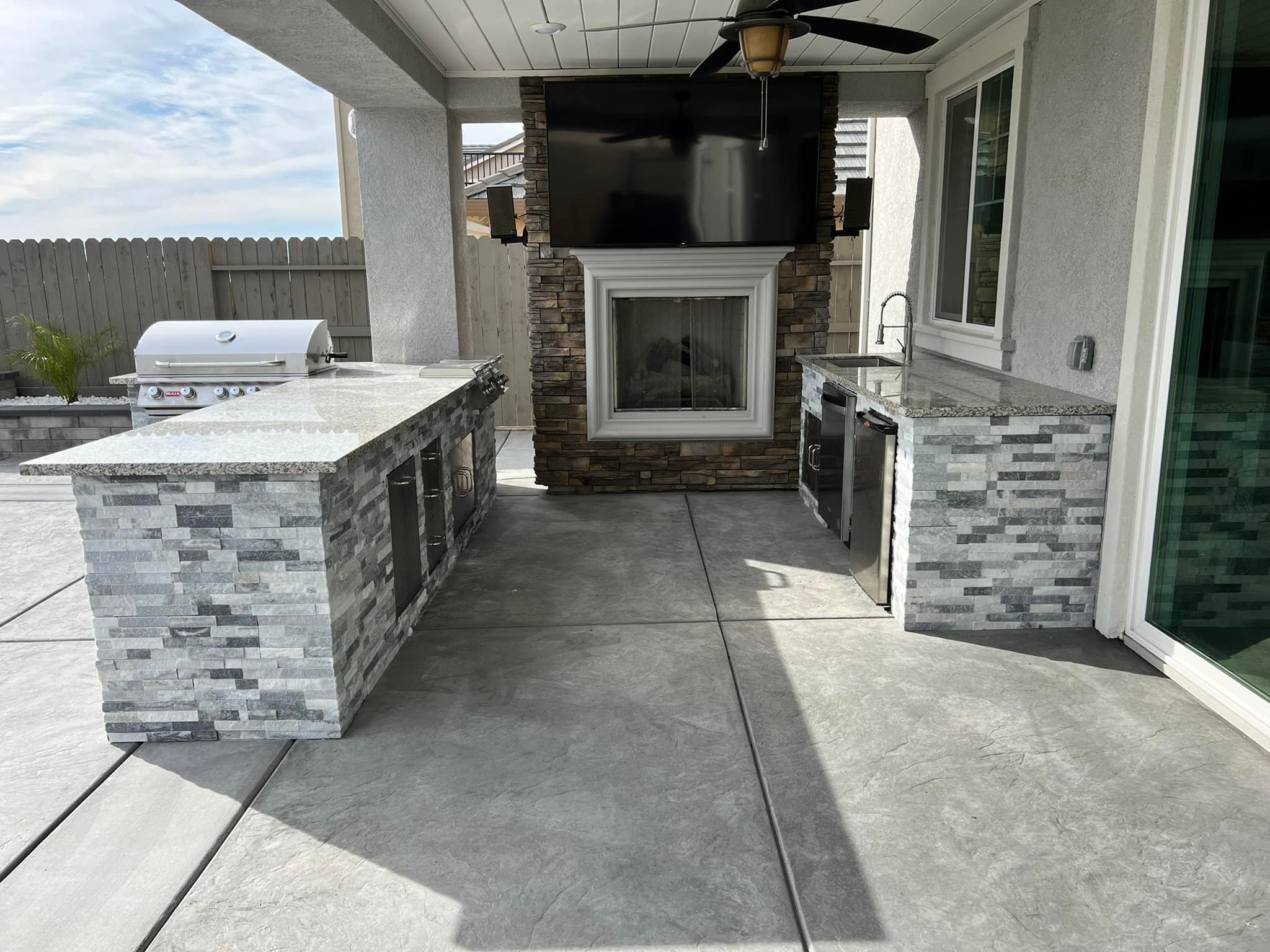 A high-end outdoor kitchen featuring premium stone masonry, granite countertops, a built-in stainless steel grill, and an outdoor fireplace under a protected patio.