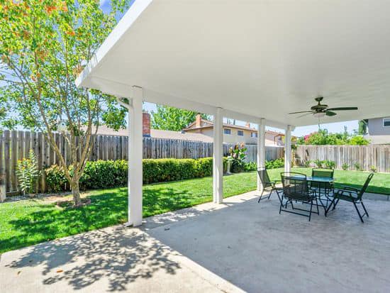 A luxury outdoor living space with a large white patio cover providing shade over a dining set on a stamped concrete floor, surrounded by green grass and mature trees.