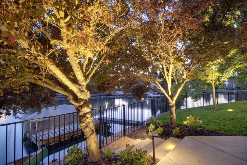 A professional residential landscaping in granite bay, CA installation featuring outdoor landscape lighting that illuminates mature trees and concrete steps leading toward a dark metal fence and a peaceful waterfront.