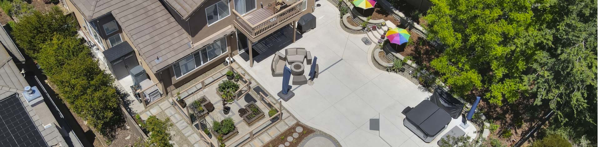 An aerial drone perspective showcasing a comprehensive, high-end residential backyard renovation. The expansive design integrates smooth concrete paving, an organized garden plot with raised wooden planter beds, a dedicated hot tub zone, and comfortable outdoor seating areas bordered by tiered retaining walls.