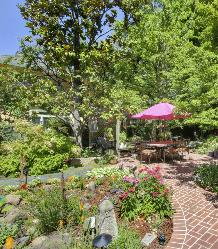 A beautifully designed backyard patio featuring a classic herringbone brick paver walkway, an outdoor dining table shaded by a vibrant pink umbrella, and a lush garden filled with pink coneflowers, mature green trees, and low-maintenance landscaping.