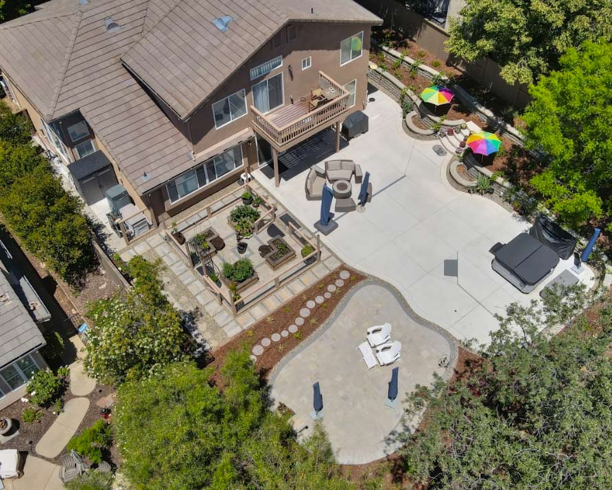 An aerial view of a complete custom backyard renovation featuring clean concrete work, custom stone patios, and an organized garden section with raised planter beds. The multi-functional layout includes a dedicated hot tub area, comfortable seating zones, and beautiful terraced retaining walls built into the slope.