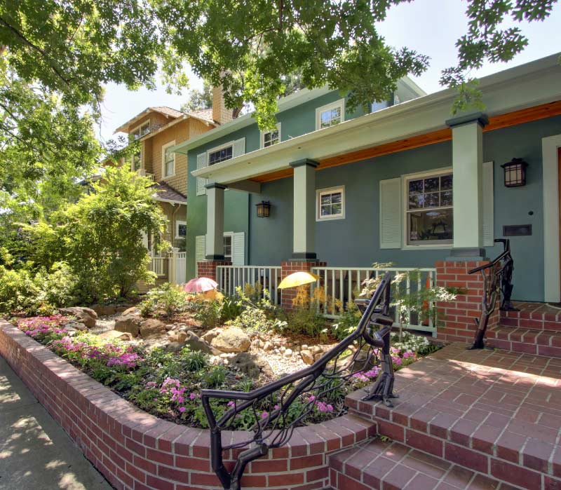 A beautifully landscaped front yard garden enclosed by a curved red brick retaining wall, featuring vibrant pink flowers, a custom rock installation, and artistic wrought-iron handrails leading up to a classic home.