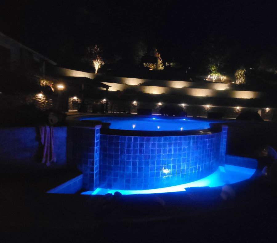 Luxury residential swimming pool at night illuminated with vibrant blue under-water accent lighting, featuring a beautifully lit multi-tiered backyard retaining wall and up-lit trees in the background.