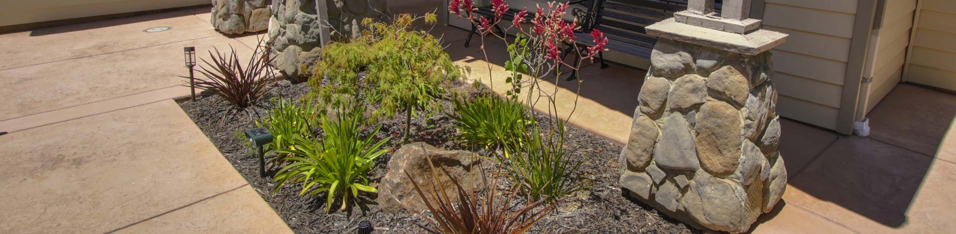 A beautiful front yard landscape display featuring clean concrete work, decorative dark mulch, and natural stone pillars. The garden bed is carefully arranged with a mix of green ornamental grasses, a small maple tree, and accent rocks that instantly boost residential curb appeal.