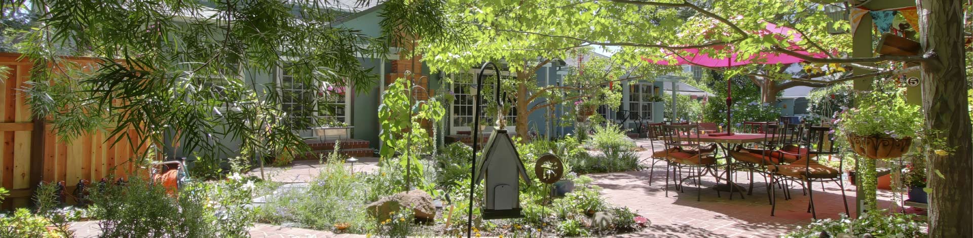 An expansive, brick-paved patio design surrounded by lush greenery, mature trees, and vibrant garden beds. The inviting outdoor dining area features a large table set under a bright pink umbrella, creating the perfect setting for relaxing entertainment.