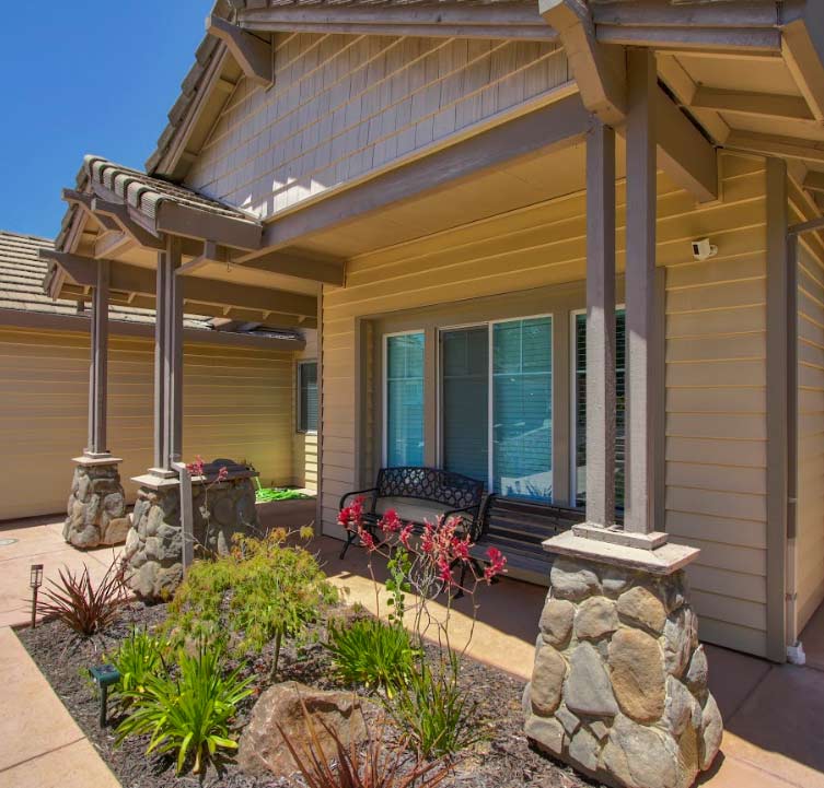 Home 1 A beautiful residential home entrance featuring a welcoming front porch, a comfortable outdoor bench, and beautifully designed pillars with a natural stone base. The front garden bed is professionally landscaped with decorative rocks and green foliage, perfectly enhancing the property's curb appeal.