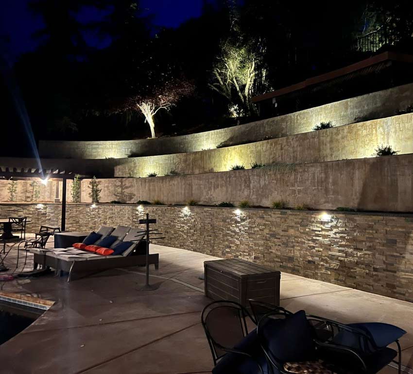 High-end outdoor landscape lighting living patio space at night showcasing a grand, multi-level stone retaining wall accented with warm spotlighting, complete with luxury poolside lounge seating.