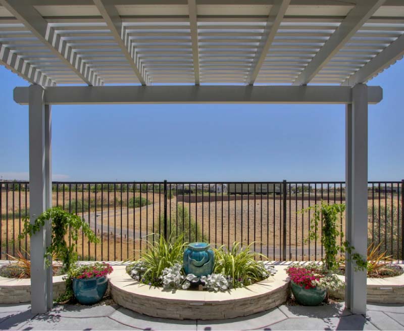 A premium outdoor living space featuring a modern white louvered pergola that provides perfect shade over a concrete patio. In the background, a custom stone planter box holds a vibrant blue ceramic water fountain surrounded by lush ornamental grasses and colorful flowers against a secure metal fence.