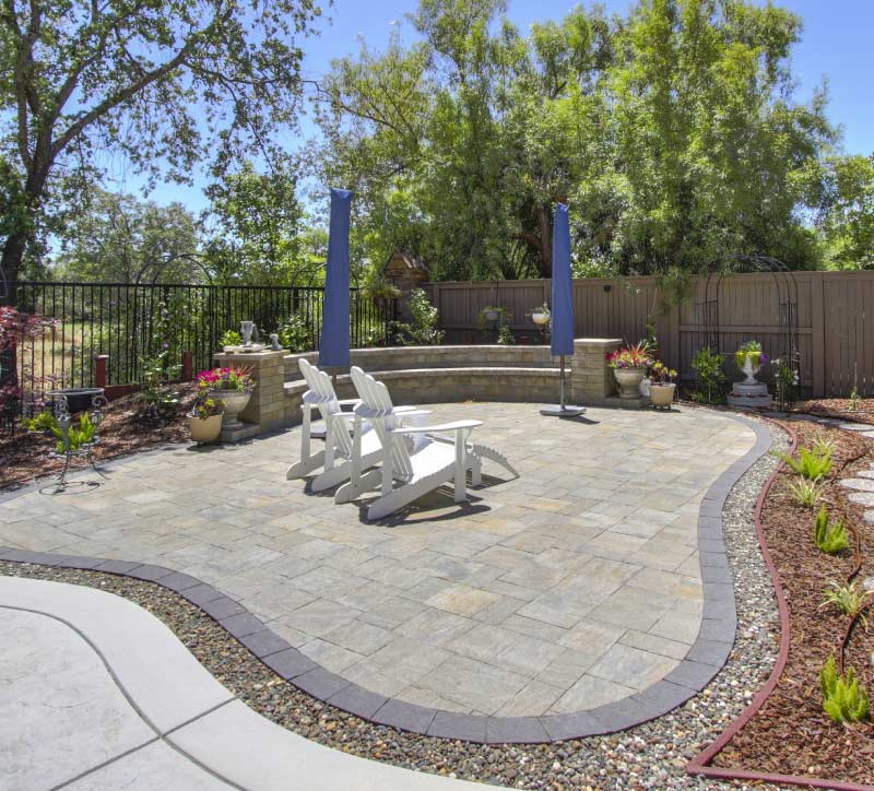 A custom-designed stone paver patio featuring a spacious seating area with white outdoor chairs, a curved stone retaining wall, and elegant landscaping that creates a perfect backyard retreat.