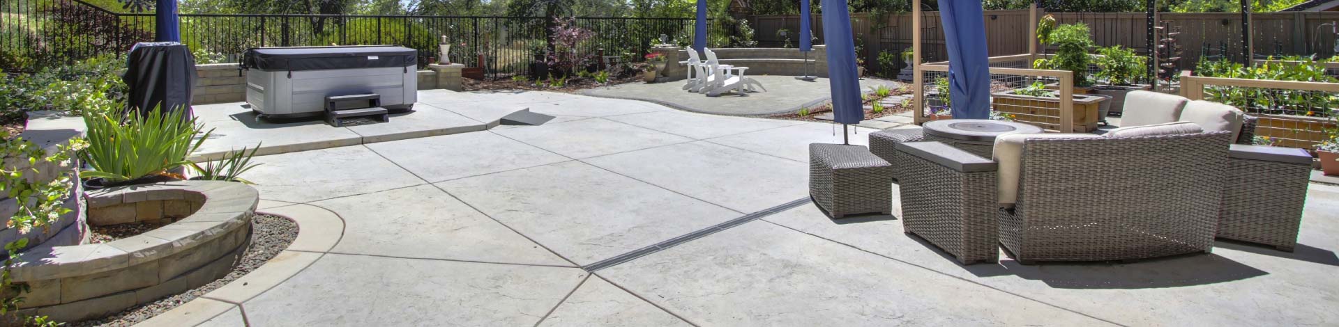 An expansive, beautifully designed concrete patio that transforms the backyard into a premium outdoor living resort. The functional design seamlessly integrates a dedicated hot tub station, an elegant wicker seating area for entertaining, and custom stone masonry features like curved planter beds and matching retaining walls.