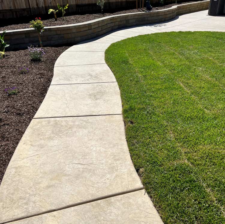 A clean, modern backyard renovation showcasing a vibrant, perfectly manicured artificial grass lawn bordered by a winding stamped concrete path and a curved stone retaining wall with fresh mulch beds.