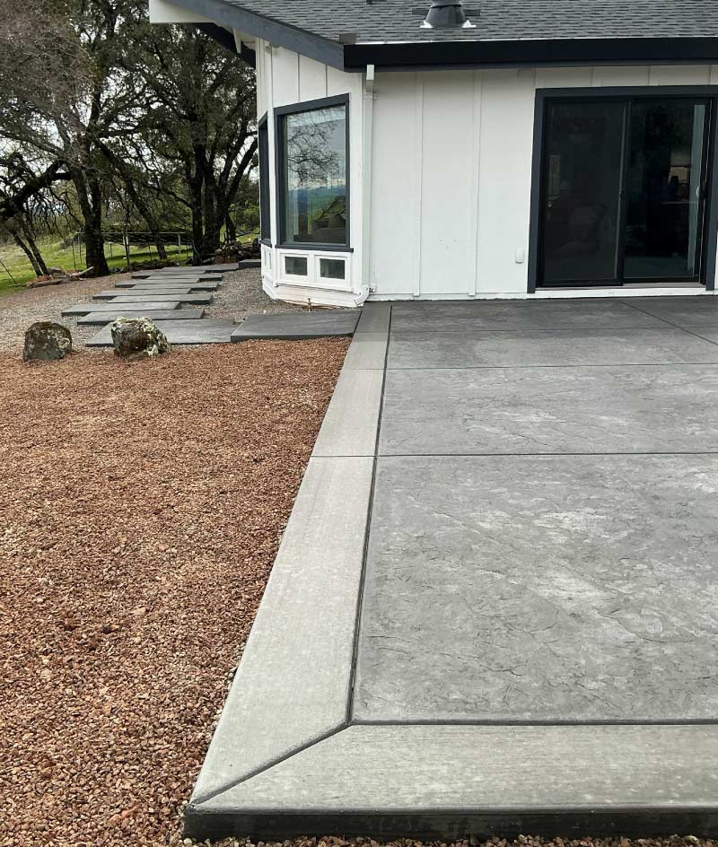 A modern backyard transformation showcasing a premium stamped concrete patio with a smooth, dark border, beautifully accented by a low-maintenance crushed rock landscape bed and large stone pavers leading to the side of the home.