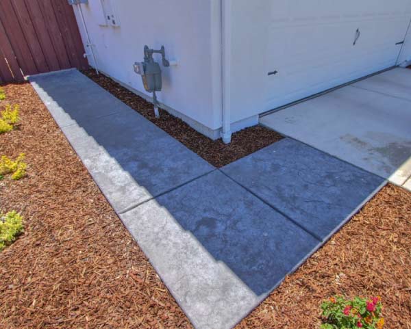Home 6 A professionally installed stamped concrete walkway with a slate-textured finish wrapping around the corner of a house. The modern charcoal gray path contrasts beautifully with fresh wood chip mulch and clean borders, highlighting low-maintenance residential landscaping.