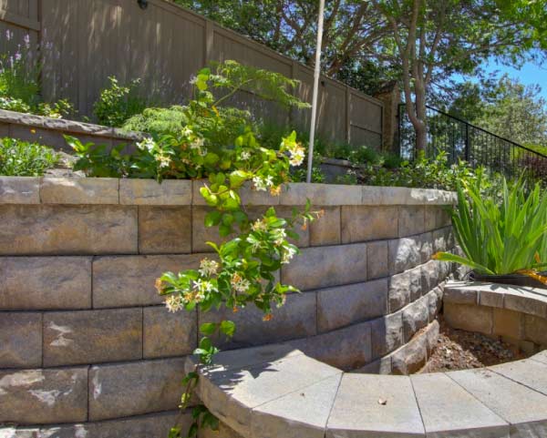 Home 4 A custom tiered retaining wall constructed with durable, multi-toned stone blocks built into a backyard slope. The structure features integrated planting beds with green foliage and delicate white climbing flowers, combining structural engineering with elegant landscape design.