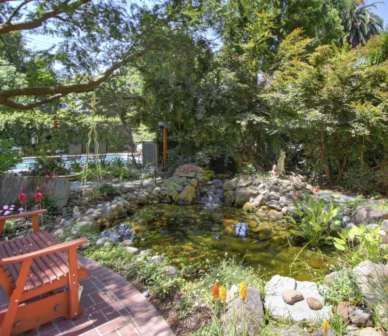 A custom backyard oasis featuring a natural rock garden pond with a cascading waterfall, nestled beneath lush green trees and a brick paver seating area.